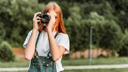 Une caméra noire est tenue devant un fond d'arbres et d'herbe verts. La caméra est au centre, devant les arbres.