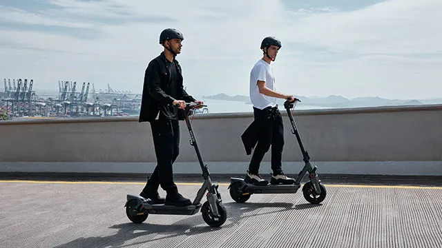Twee personen op grijze elektrische scooters langs een grijze muur met op de achtergrond een haven en een blauwe lucht.