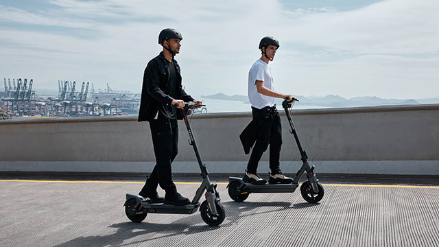 Deux personnes sur des scooters électriques gris, portant des casques, sur une route grise avec une rambarde. En arrière-plan, des grues portuaires et un ciel nuageux.