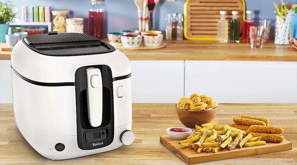 A white deep fryer sits near a wooden board with fries, sticks, and sauce. A bowl of onion rings is also present.