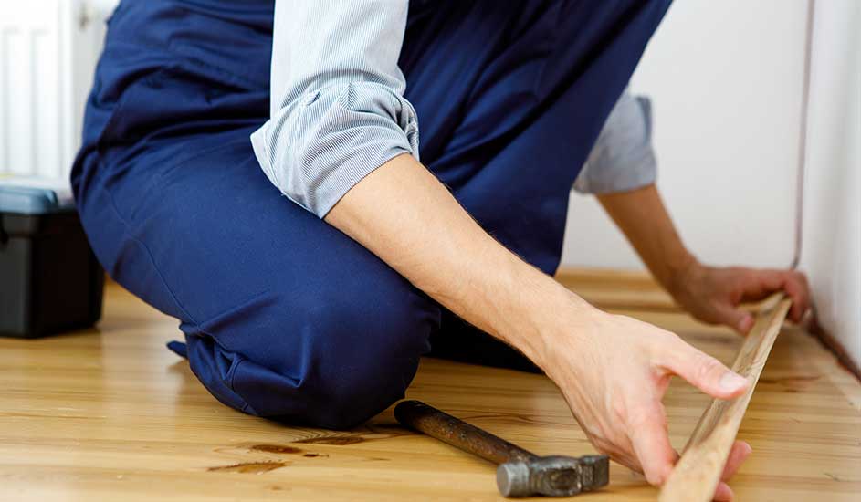 Un ouvrier en bleu pose une plinthe en bois sur un plancher. Un marteau et une boîte à outils sont visibles.