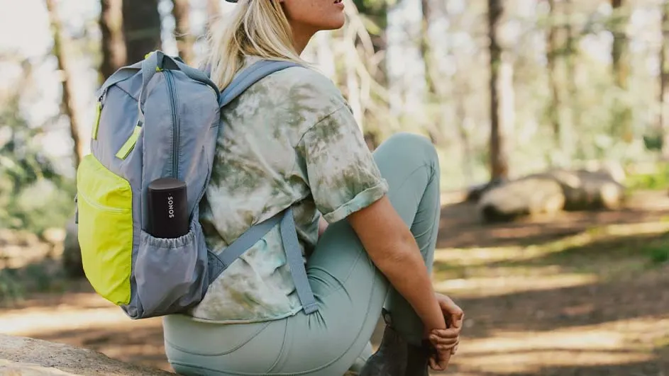 Une personne assise avec un sac à dos gris et jaune, contenant un haut-parleur noir Sonos, dans un environnement forestier.