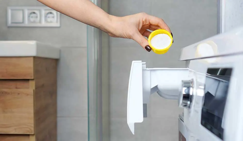 Hand giet wit wasmiddel uit een gele dop in een witte wasmachine. Achtergrond: grijze muur, houten kast.