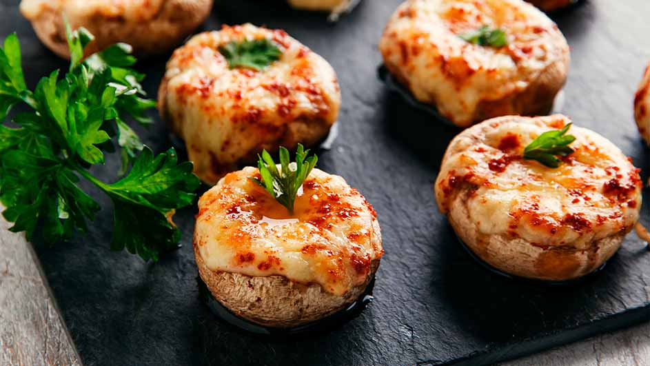 Des champignons farcis au fromage, cuits et garnis de persil, sont présentés sur une plaque noire.