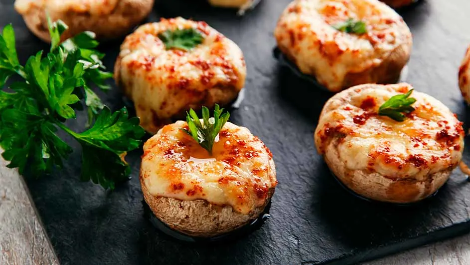 Des champignons farcis au fromage, cuits et garnis de persil, sont présentés sur une plaque noire.