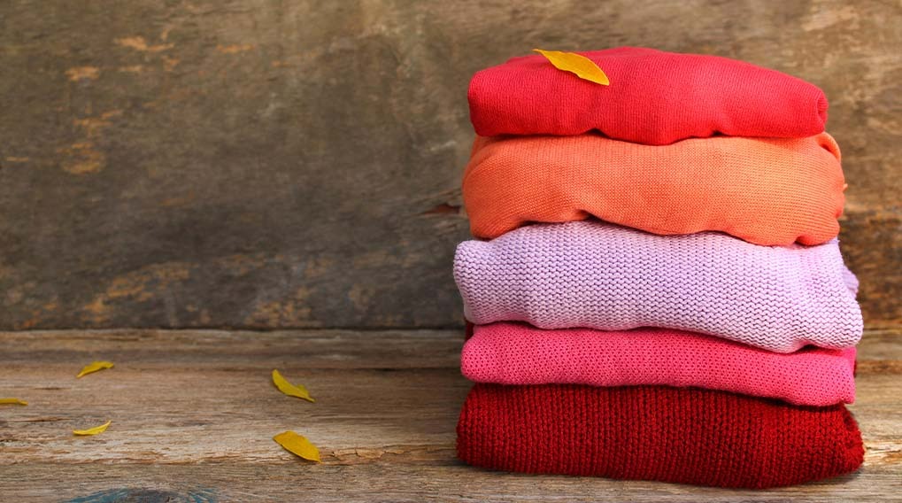 Un tas de pulls tricotés colorés (rouge, orange, lilas, rose, rouge foncé) sur du bois avec des feuilles jaunes éparses.