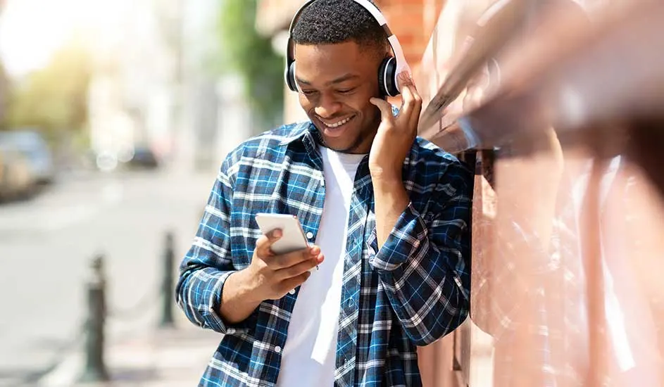 Un homme avec un casque blanc tient un smartphone. Il porte une chemise à carreaux bleue et blanche et un t-shirt blanc.