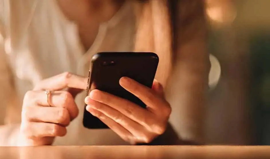 Een persoon in een lichte blouse gebruikt een zwarte smartphone met één hand, zittend aan een houten tafel in warm licht.