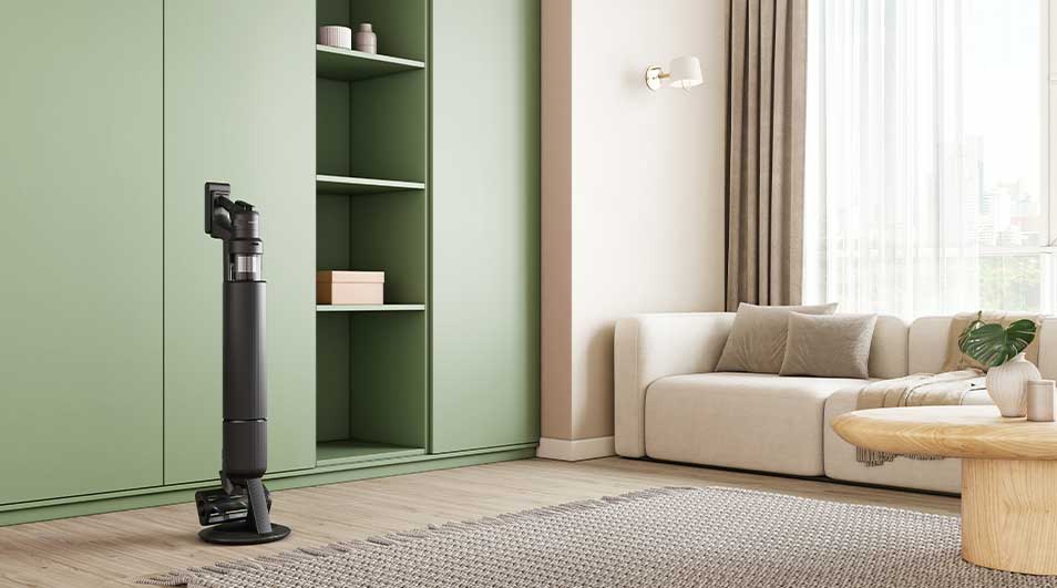 Aspirateur balai noir posé sur base ronde devant grand placard vert, dans un salon chaleureux avec canapé beige et table en bois.