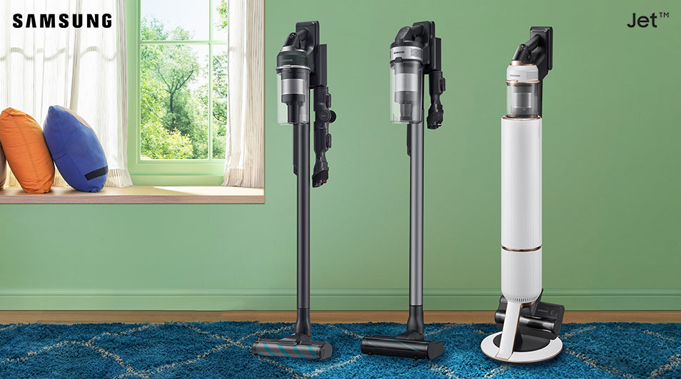 Trois aspirateurs verticaux Samsung, noir, gris et blanc, sur un tapis bleu, devant un mur vert clair et une fenêtre.