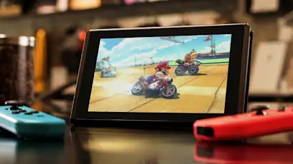 A Nintendo Switch is on a table, showing Mario Kart gameplay onscreen, with red and blue Joy-Con controllers placed on each side.