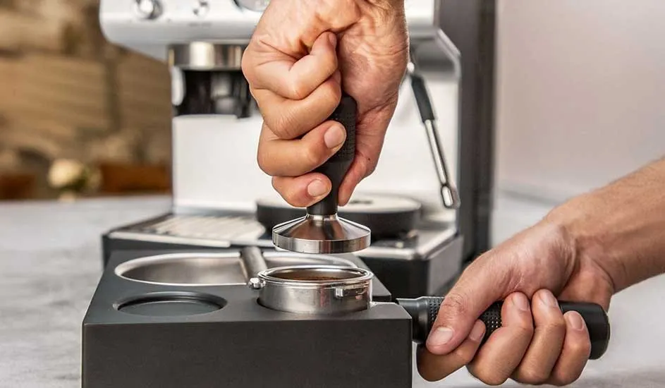 Hands firmly tamp fresh espresso grounds in a metal portafilter on a tidy station, with a modern coffee machine waiting in the background.
