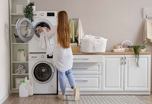 Een wasmachine staat open in een wasruimte met witte kasten en een wasmand. Er staat wasmiddel op een plank.