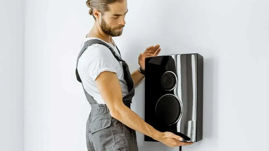 A black speaker with two round openings is mounted on a white wall. Part of a person in grey overalls.