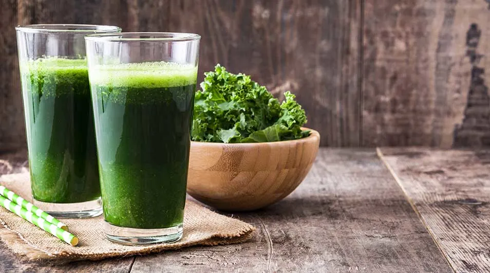 Deux verres de jus vert, un bol de chou frisé, des pailles et une nappe sur une table en bois.