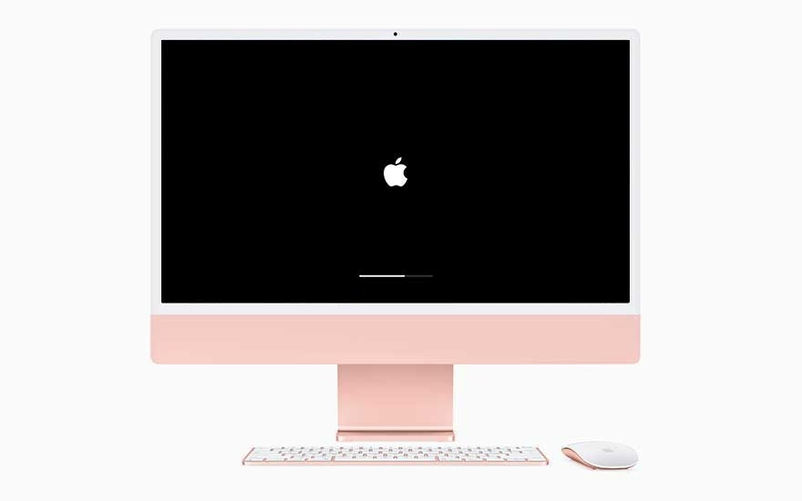 A pink iMac on a white surface with a black screen displays a white Apple logo and loading bar. A white keyboard and mouse are below.