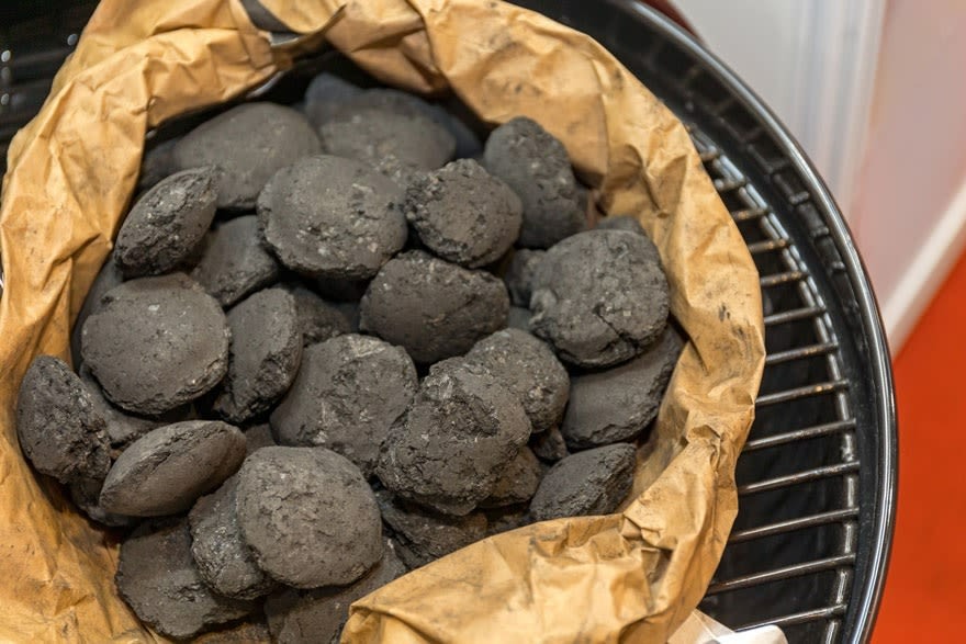 Dans un sac en papier brun, des briquettes de charbon gris reposent sur une grille de barbecue noire.