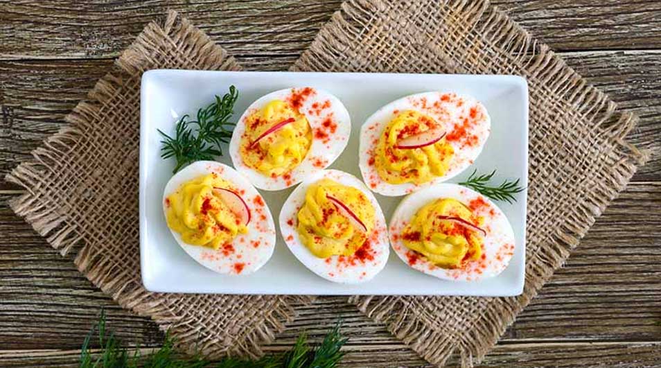 Vijf gevulde eieren op een wit bord, met paprika en peterselie, op een jute onderzetter op een houten tafel.