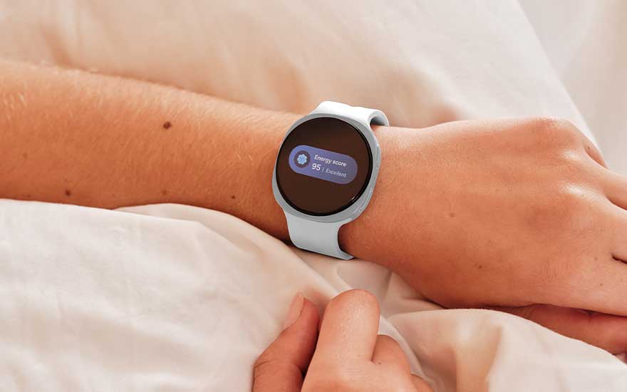 Een ronde  Galaxy Watch9 met lichtblauwe band toont een energyscore op een pols die rust op zachte witte lakens.