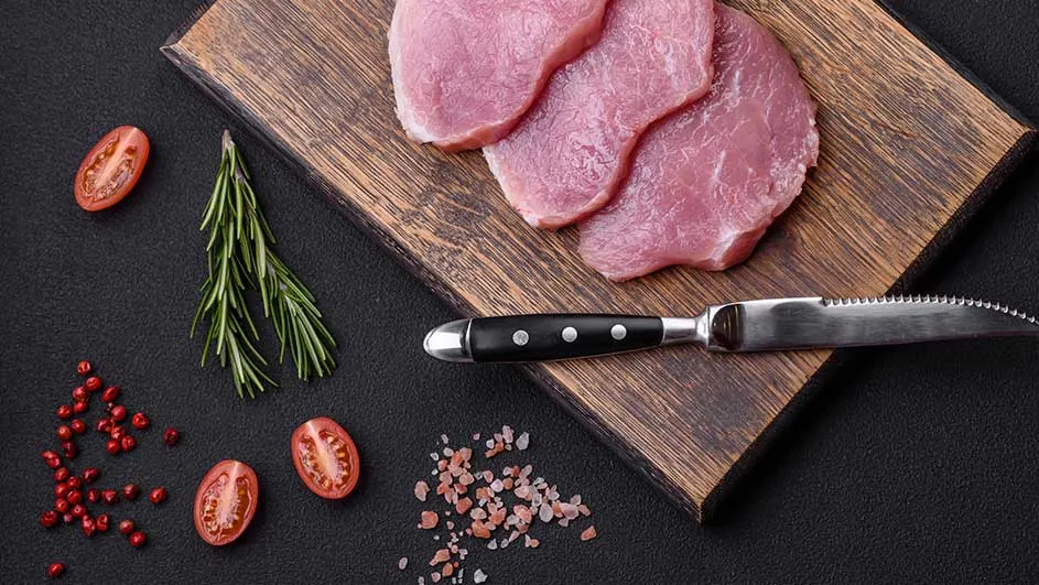 Sur une planche à découper en bois se trouvent des tranches de viande crue, un couteau, des tomates cerises, du romarin et du sel rose sur un fond noir.