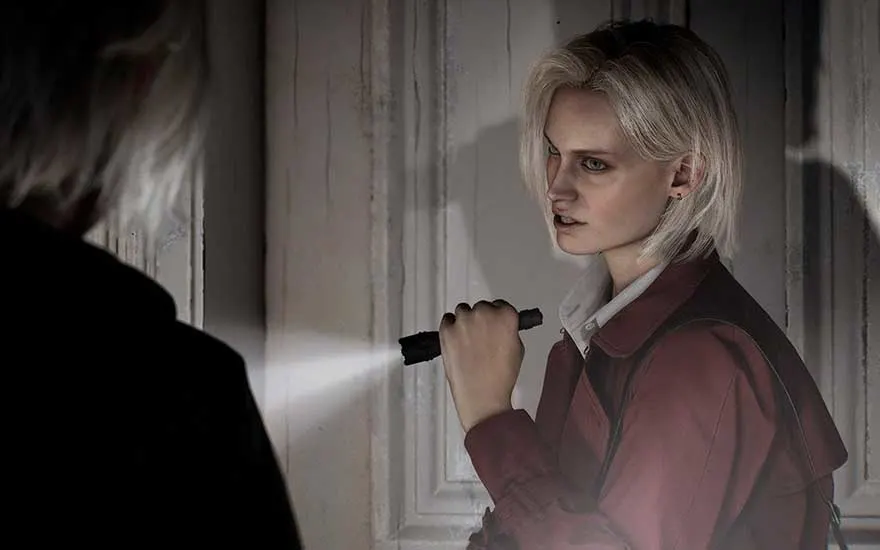 A young blonde woman tensely inspects a peeling old door, shining her torch across the wood while a shadowy figure stands nearby.