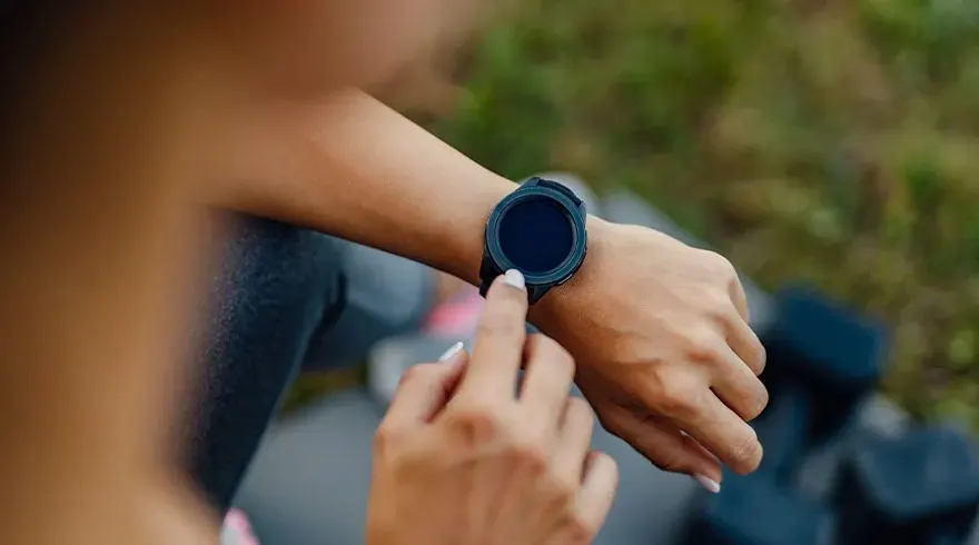 Une montre noire est au poignet, un doigt touche l'écran. L'arrière-plan est flou, avec des poids noirs.