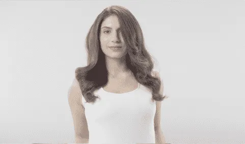 Une personne aux cheveux longs ondulés et châtains porte un débardeur blanc devant un fond blanc.