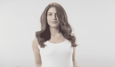 Une personne aux cheveux longs ondulés et châtains porte un débardeur blanc devant un fond blanc.