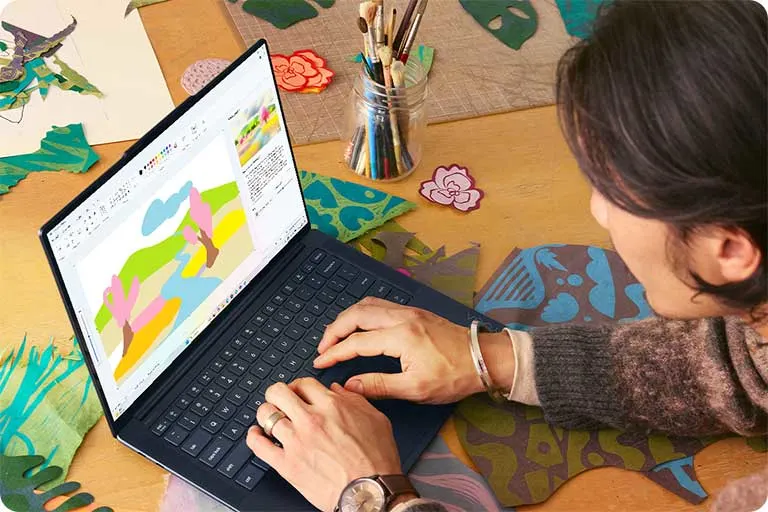 Een laptop toont een kleurrijk digitaal schilderij op een houten tafel met verfborstels, papieren bloemen en uitgesneden vormen.