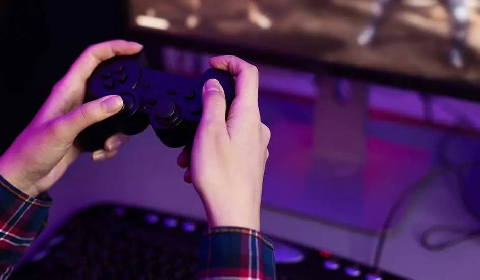 Deux mains tiennent une manette noire Sony PlayStation devant un écran de jeu, dans une pièce éclairée en violet.