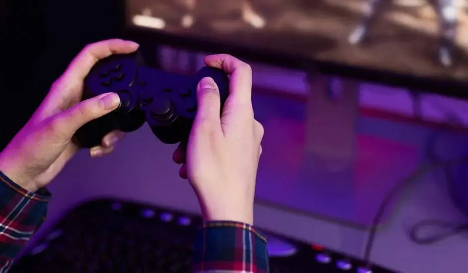Deux mains tiennent une manette noire Sony PlayStation devant un écran de jeu, dans une pièce éclairée en violet.