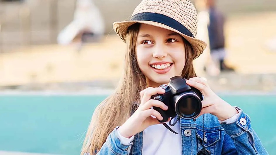 Une personne tient un appareil photo noir devant un fond bleu. Elle porte un chapeau de paille.