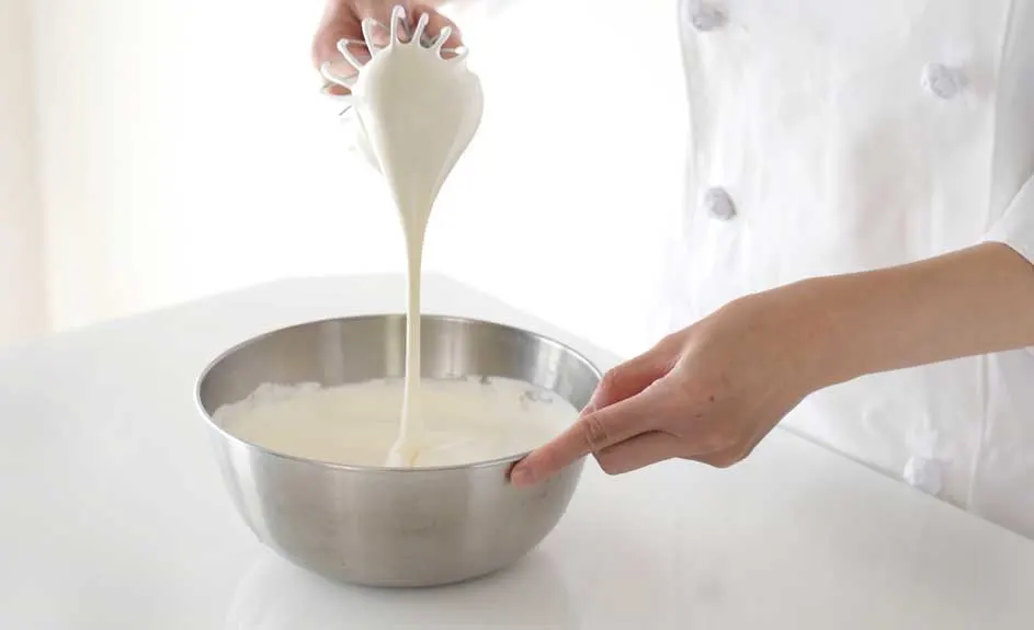 Un chef en blouse blanche verse une pâte claire d'un fouet dans un bol en métal sur une table blanche.