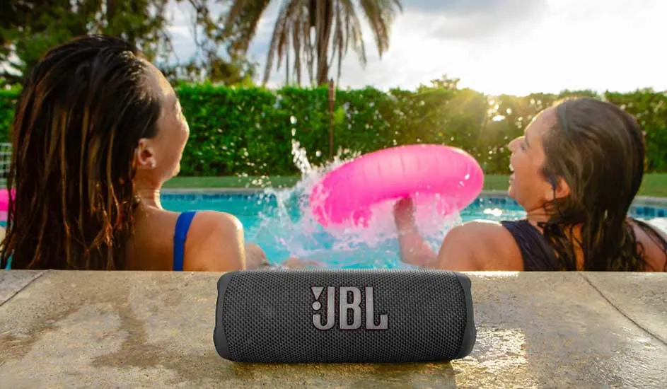 Deux personnes dans une piscine jouent avec une bouée rose. Un haut-parleur JBL gris est au premier plan.