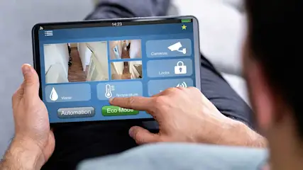 Un homme tient une tablette noire avec un écran bleu affichant des contrôles pour la maison, tels que la température et les caméras.