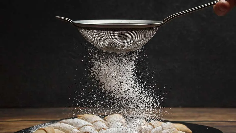 Een metalen zeef strooit witte poedersuiker over gebak op een zwarte schaal, tegen een donkere achtergrond.