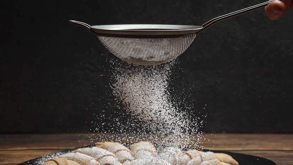 Un tamis verse du sucre glace sur des croissants. Les croissants sont sur une assiette noire, sur une table en bois. Fond noir.