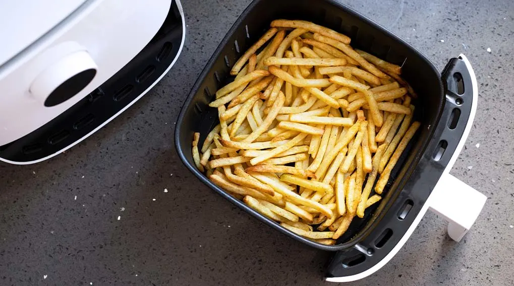 A black air fryer basket is filled with golden fries, next to a white air fryer on a speckled countertop.