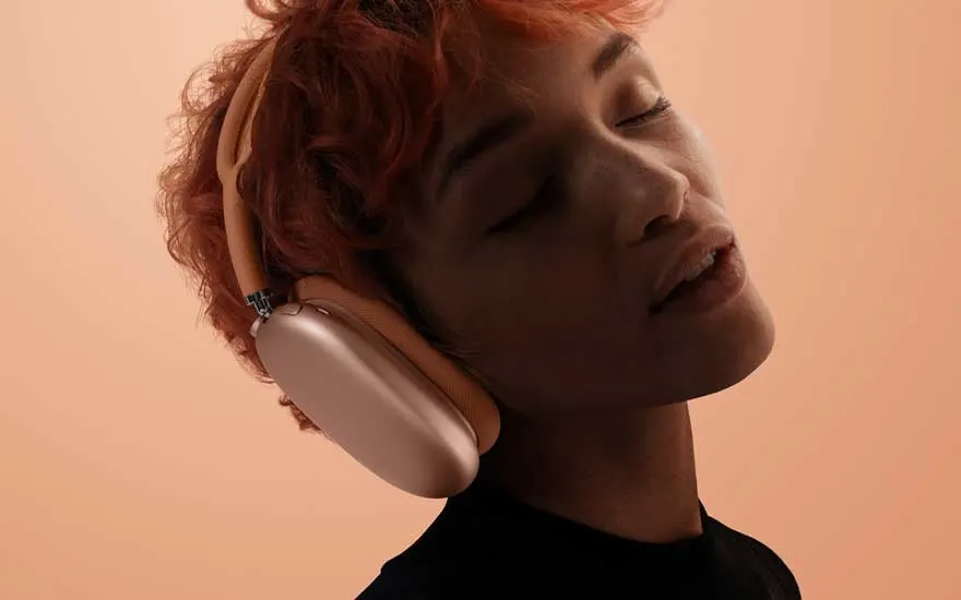 Een persoon met kort oranje haar draagt perzikkleurige over-ear hoofdtelefoon en sluit genietend de ogen voor een zachte abrikooskleurige achtergrond.