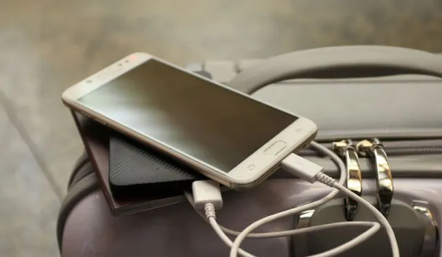 Een witte smartphone ligt op een zwarte powerbank op een grijze koffer, aangesloten met een witte kabel.