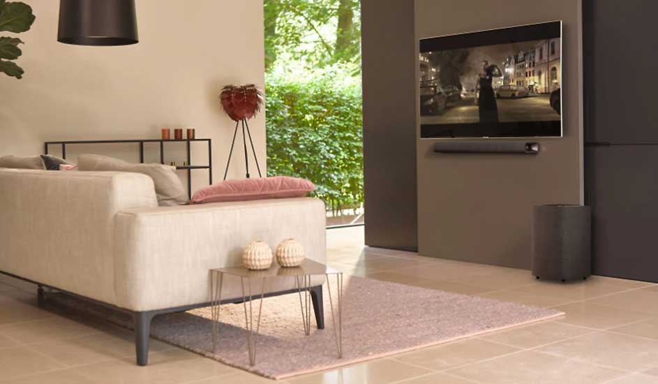 Een woonkamer toont een beige sofa met kussen, een salontafel met decoratie, en een tv aan de muur. Buiten is groen te zien.