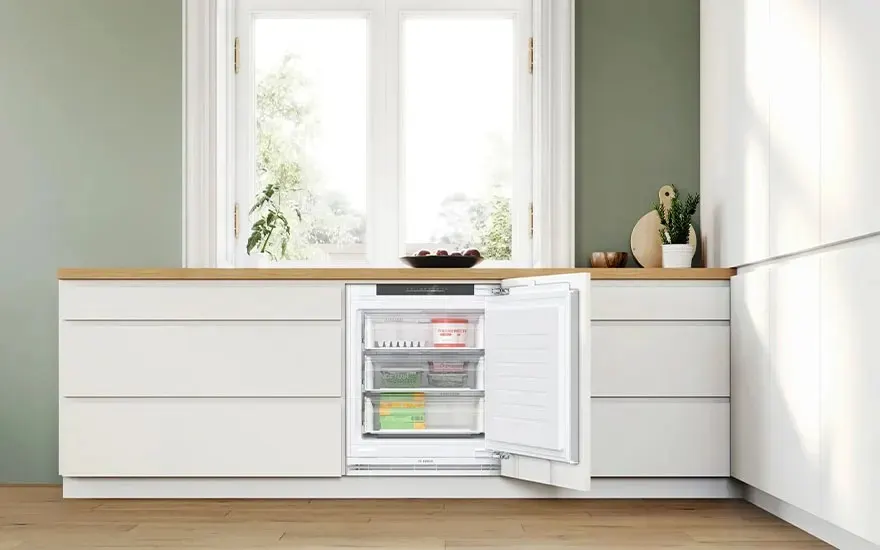 Un congélateur encastrable Bosch porte ouverte, trois tiroirs transparents remplis d’aliments, intégré dans une cuisine blanche moderne sous une fenêtre.