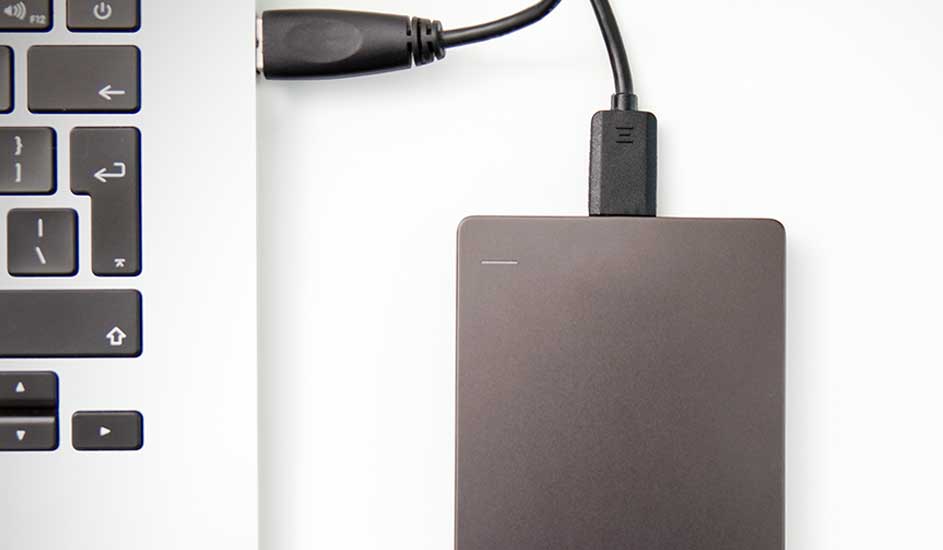 Un disque dur externe gris est connecté à un ordinateur portable argenté par un câble USB noir sur un fond blanc.