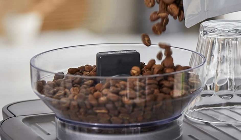 Brown coffee beans tumble into a clear grinder hopper, filling the transparent bowl above a sleek machine, ready for a fresh brew