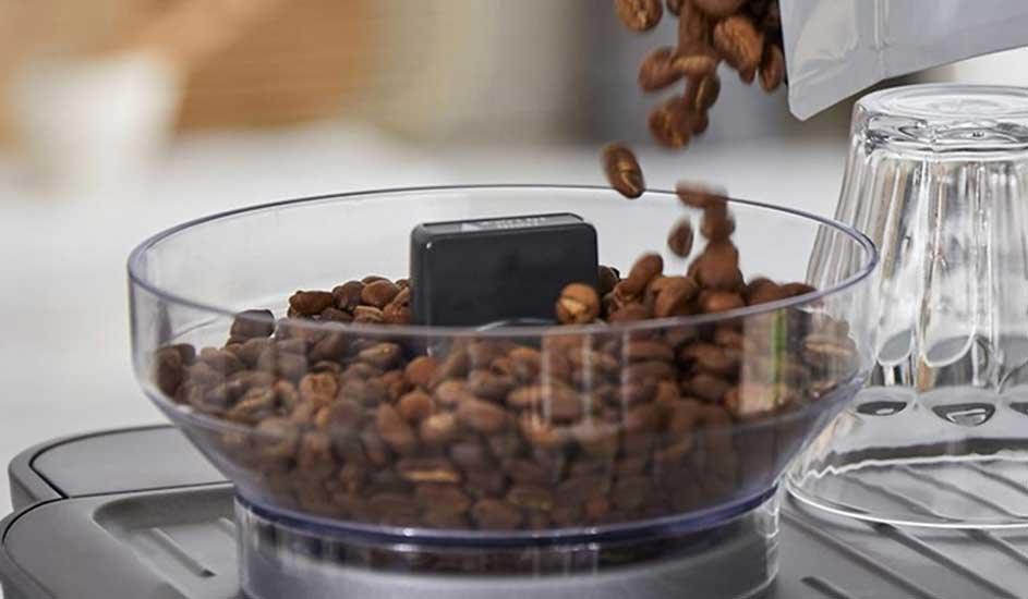 Des grains de café tombent d’un sachet dans un réservoir transparent posé sur la machine, à côté de verres retournés.