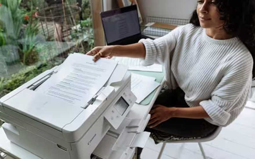 Een vrouw haalt een geprint document uit een witte printer. Ze werkt thuis aan een tafel met een laptop en andere papieren naast zich.