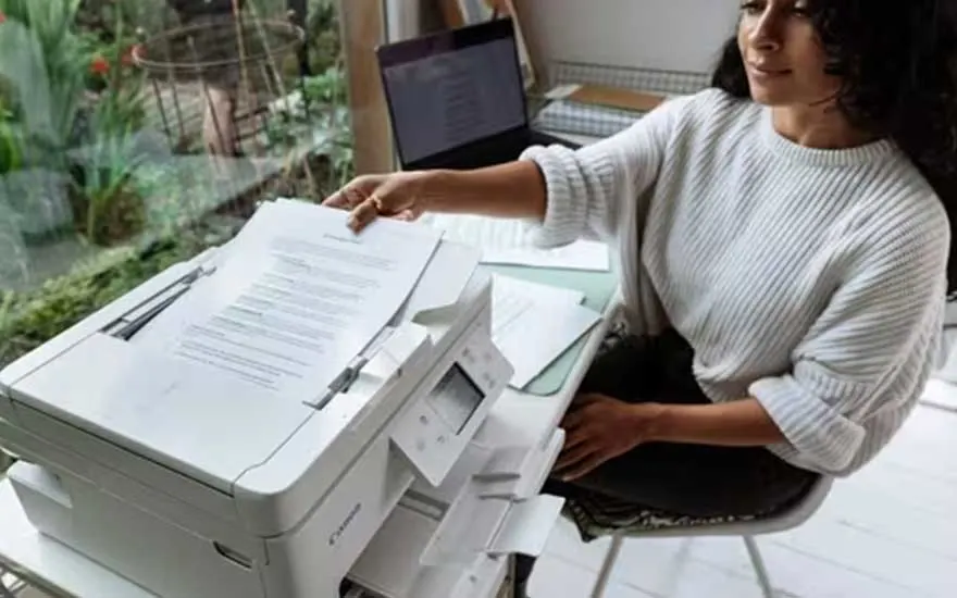 Een vrouw haalt een geprint document uit een witte printer. Ze werkt thuis aan een tafel met een laptop en andere papieren naast zich.