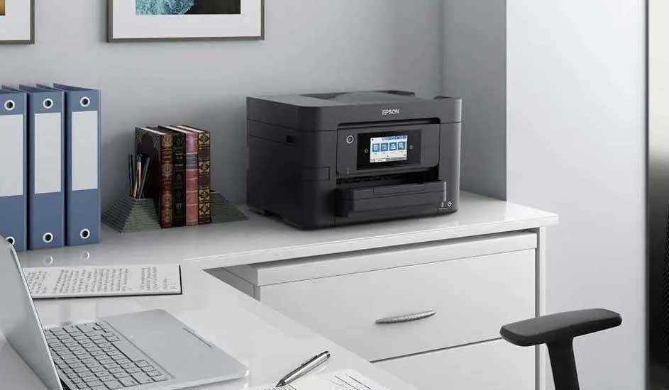 Bureau blanc avec imprimante noire, ordinateur portable et classeurs bleus. Document et stylo visibles.