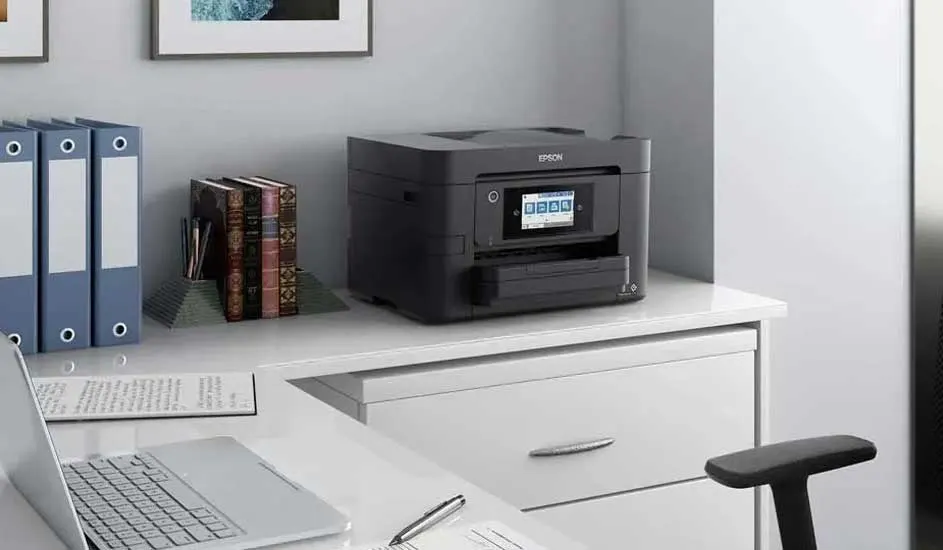 Bureau met laptop, printer, ringmappen en boeken. De printer staat op een witte ladekast tegen een grijze muur.