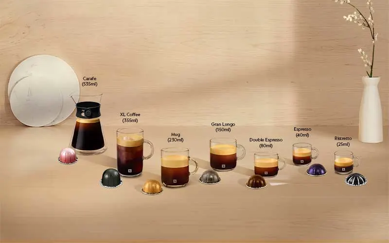 Op een houten tafel staan zeven soorten Nespresso koffie, van carafe tot ristretto, met bijbehorende cups en een witte vaas rechts.
